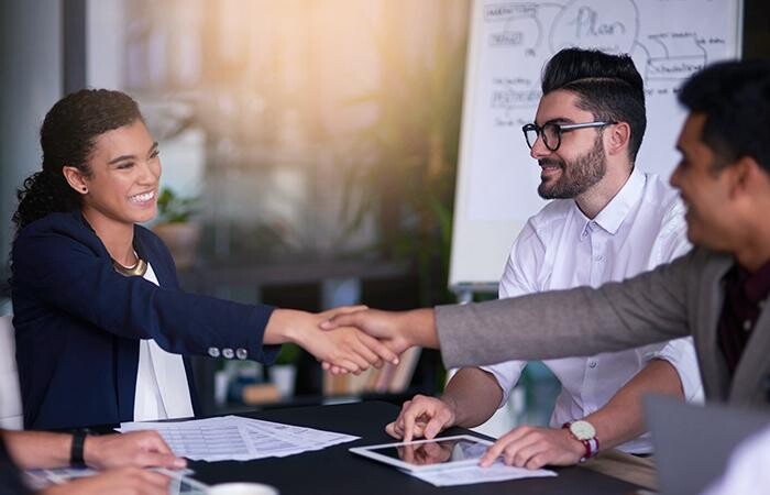 Three workers collaborating around desk, with a pair shaking hands, confirming their marketing ideas.